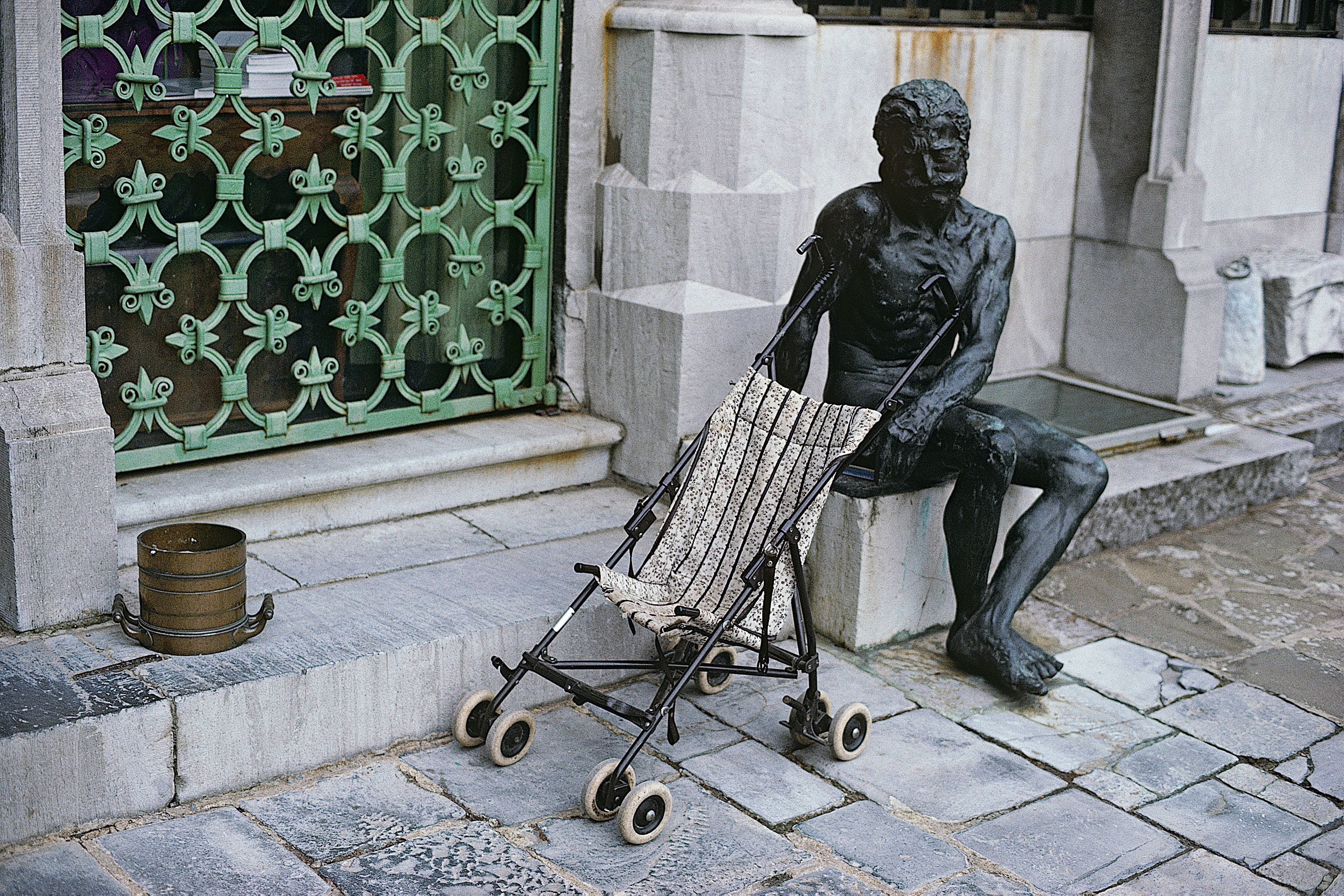 Bronzeskulptur eines sitzenden Mannes, davor ein leerer Kinderbuggy vor grünem Ziergitter