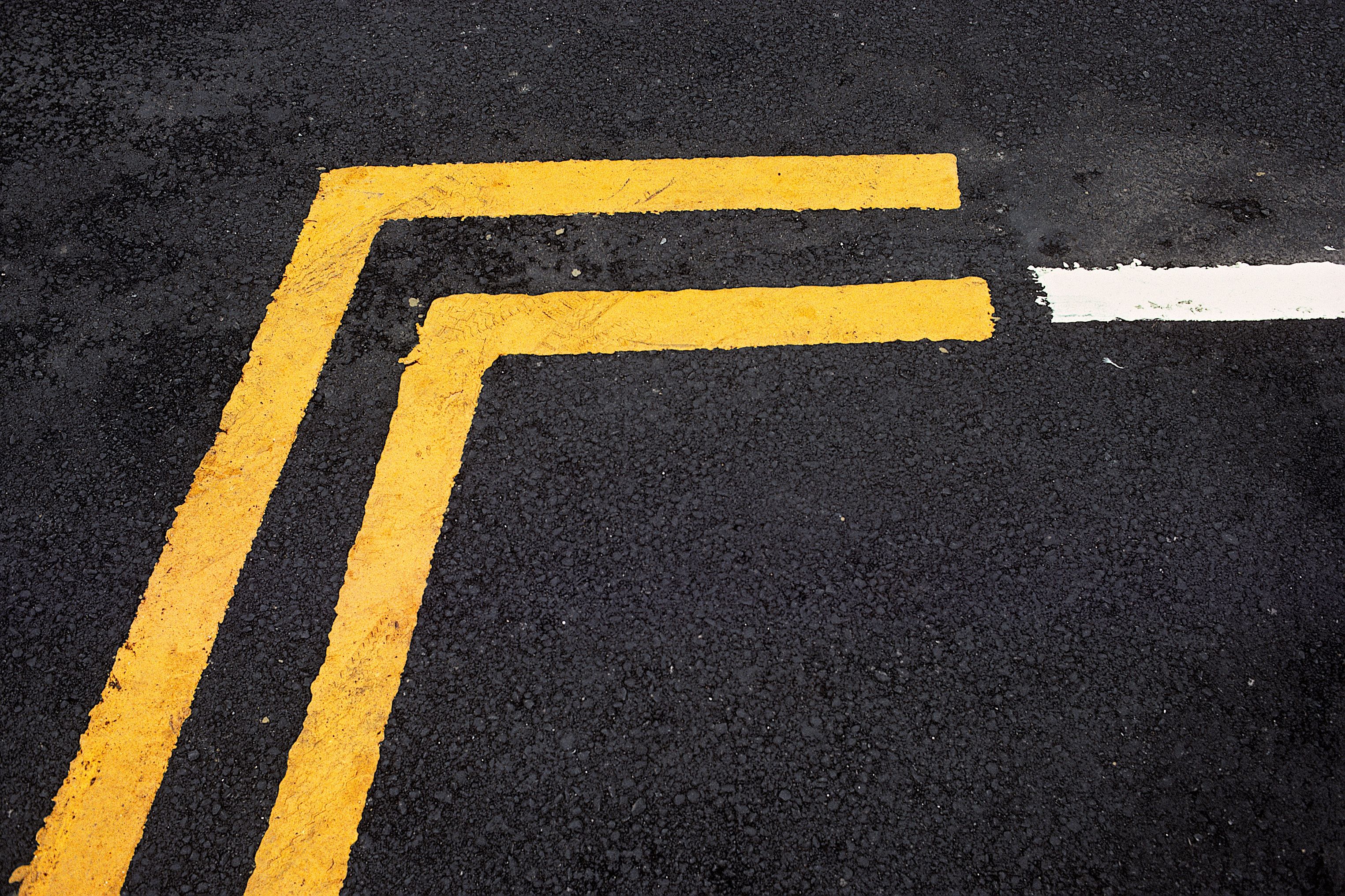 Gelbe Fahrbahnmarkierungen im rechten Winkel auf schwarzem Asphalt, rechts eine weiße Linie